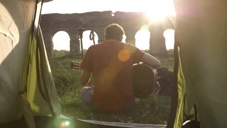 young man backpacker playing guitar outside camping tent sitting on grass in front of roman aqueduct arches in parco degli acquedotti park ruins in rome at sunset sticks in ground camera shot inside tent pov slow motion