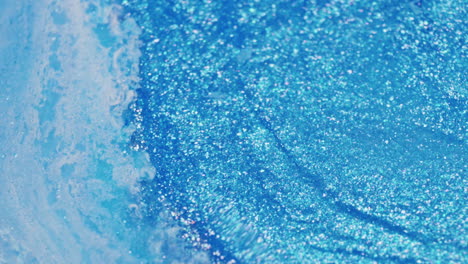 macro shot of glittery blue ink rippling and swirling as a water droplet lands off-frame, creating surface movement and waves