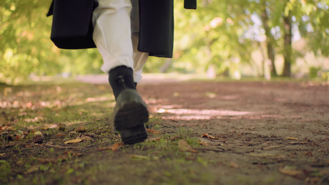 Leg view of carefree walker in black boots, white pants, long black coat, wandering under pine grove on shaded path, slow steps over leaf carpet, soft bokeh, nature walk with fallen leaves on ground