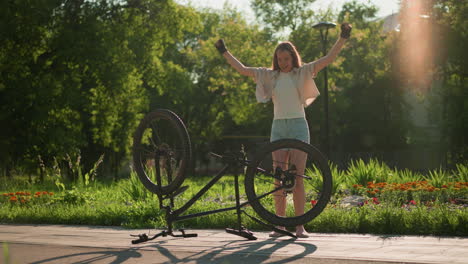 young lady joyfully jumping and dancing next to her upside-down bike, expressing happiness and excitement in a sunlit outdoor setting, with blurred greenery creating a vibrant backdrop