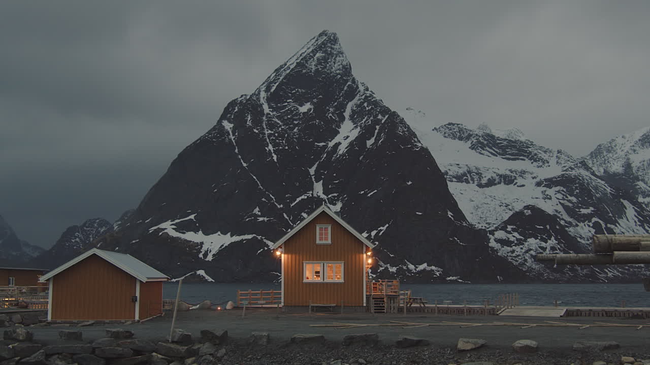 Vista pitoresca de uma pequena cabana iluminada com a montanha Olstinden ao fundo ao crepúsculo na vila de Sakrisoy, Ilhas Lofoten, Noruega - plano aberto