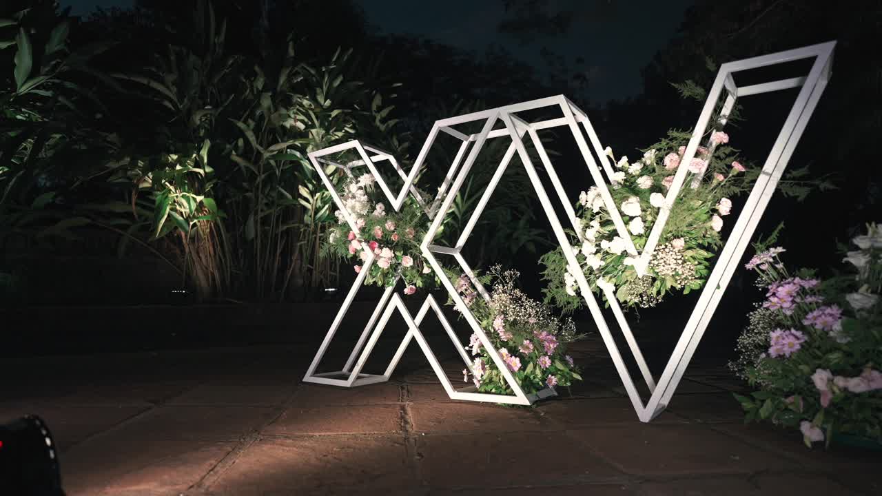 Illuminated metal letters forming “XV” stand on garden path at night, with open geometric frames filled with pastel flowers, creating modern decorative backdrop for Sweet 15 quinceañera celebration
