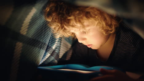 happy little boy using digital tablet computer under blanket enjoying learning on touchscreen technology playing games having fun at bedtime