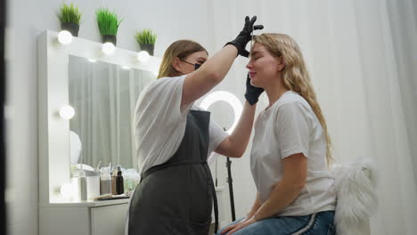 beautician in white top and apron uses picker to groom client eyebrow in well-lit salon. client sits calmly on fluffy chair wearing white top and jeans, reflecting clean modern beauty care environment