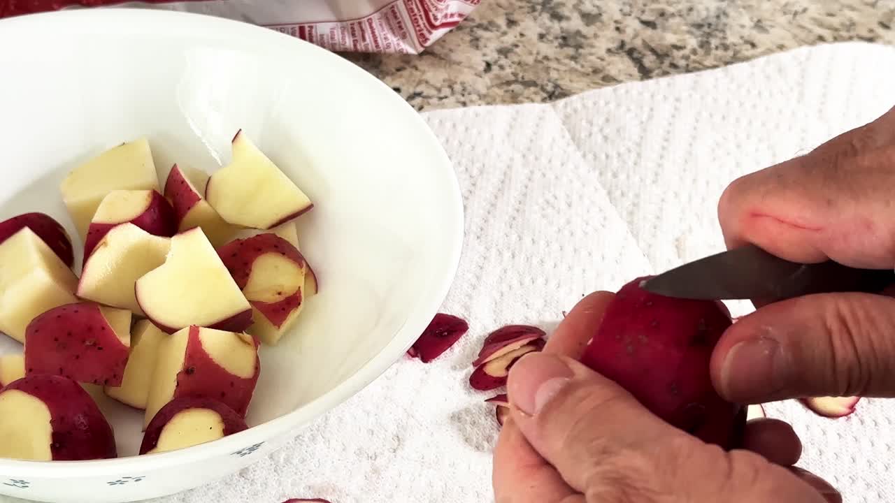 Close-up view of male hands using a paring knife to peel and chop small red potatoes. The pieces are collected in a white bowl for meal prep