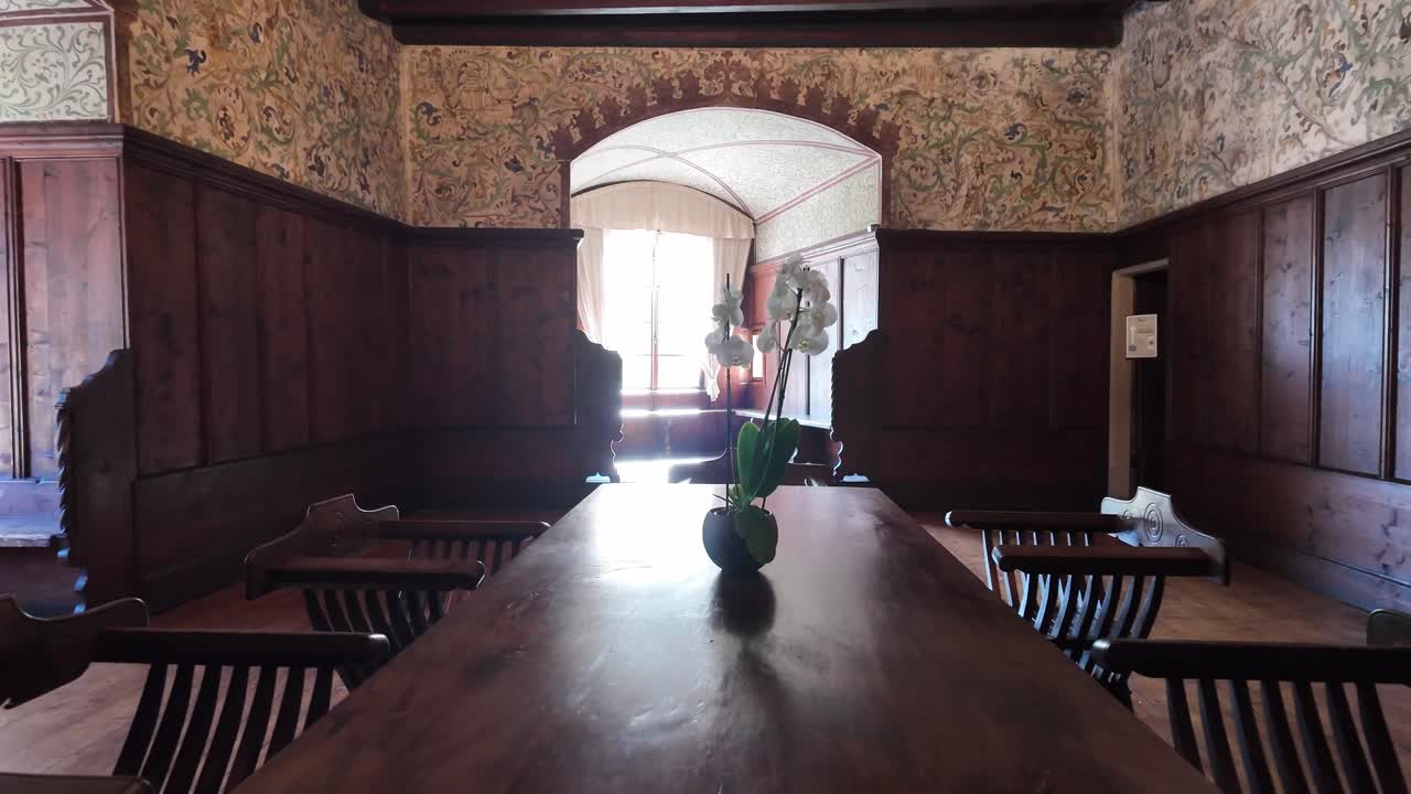 A beautifully decorated castle chamber with richly decorated walls, large wooden table, surrounded by carved chairs in the center stands. White flowers stand in the center of the table