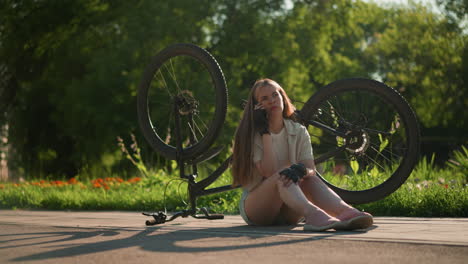 young lady seated on the pavement beside her upside-down bike, answering phone call with a concerned look, surrounded by lush greenery and trees in outdoor park setting