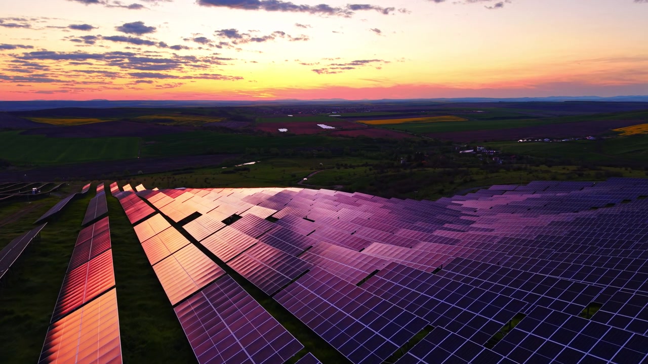 Fotovoltaisk solcellepark ved skumringstid. Vidvinkel oversigtsbillede af massivt bæredygtigt energianlæg, der reflekterer rød solnedgangsglød nær mørkt bølgende mark.