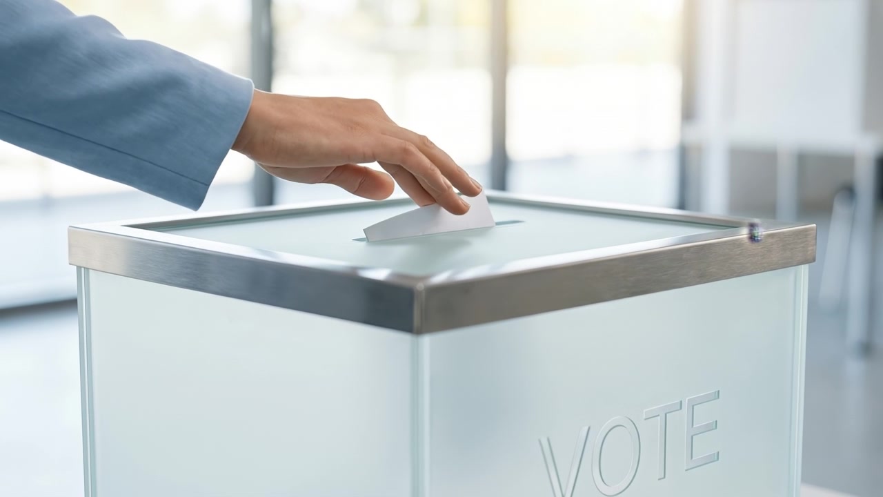 Close up of a hand inserting a white ballot paper into a ballot box with vote text