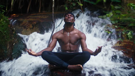 black young male model sits in meditative yoga pose in a waterfall with legs crossed and eyes closed, hands in chin mudra, breathing deeply, water flowing in rainforest environment