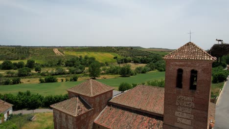 Campanario-De-Ladrillo-De-La-Iglesia-Con-Nido-De-Cigüeñas-Y-Verdes-Tierras-De-Cultivo-En-El-Fondo