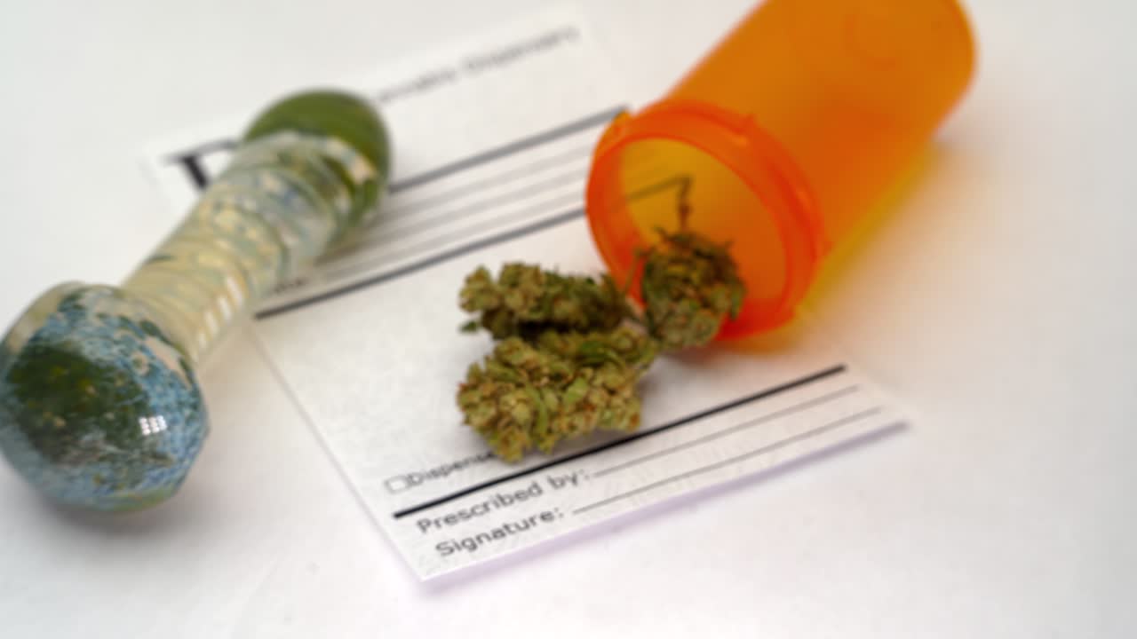 foco de rack de una pipa de marihuana de vidrio, botella de prescripción abierta, flores de marihuana y nota rx en blanco sobre una mesa blanca