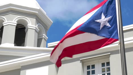 Die-Puerto-ricanische-Flagge-Weht-Im-Wind,-Während-Eine-Taube-Aus-Dem-Turm-Eines-Gebäudes-Fliegt