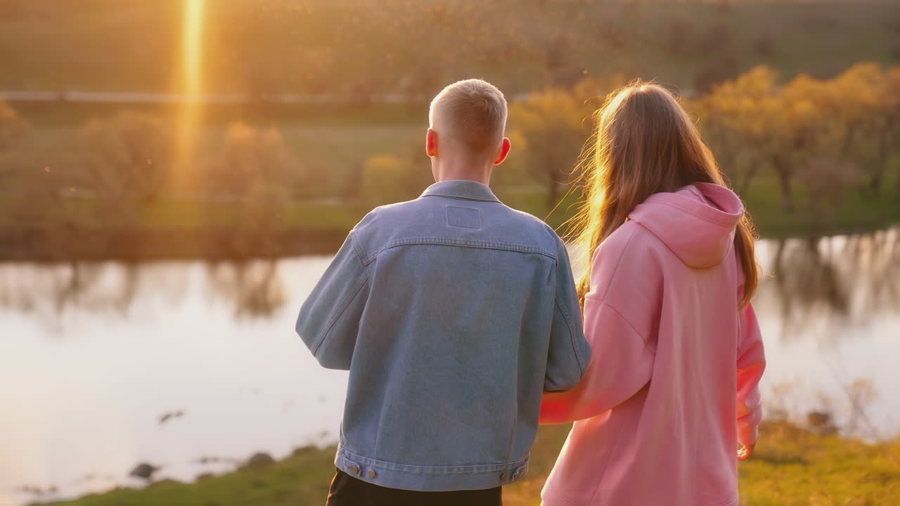 Romantic couple at sunset. Beautiful girl and her boyfriend standing in nature and looking at the setting sun. Happy young people talk near the river.