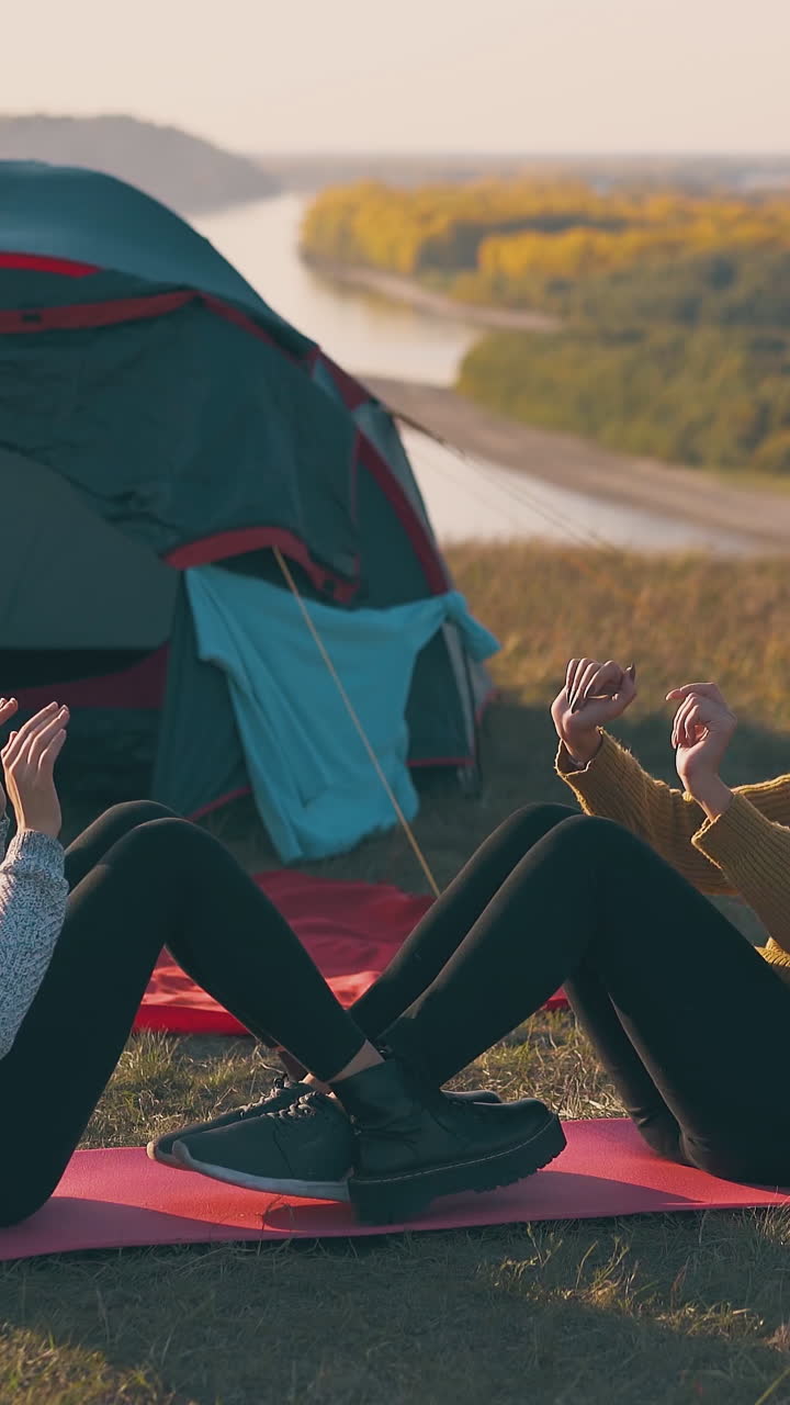 joyful girlfriend tourists do abs exercises on mat at comfortable campsite with tent at sunset in autumn evening
