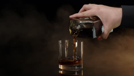 barman pour whiskey cognac brandy from bottle into drinking glass with ice cubes on dark background