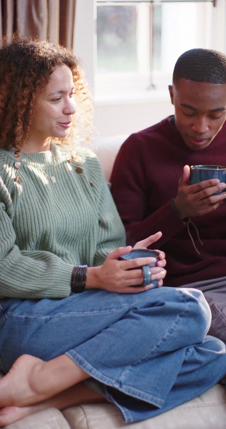 Vertical video: Diverse couple smiling and catching up on couch in living room, holding blue mugs