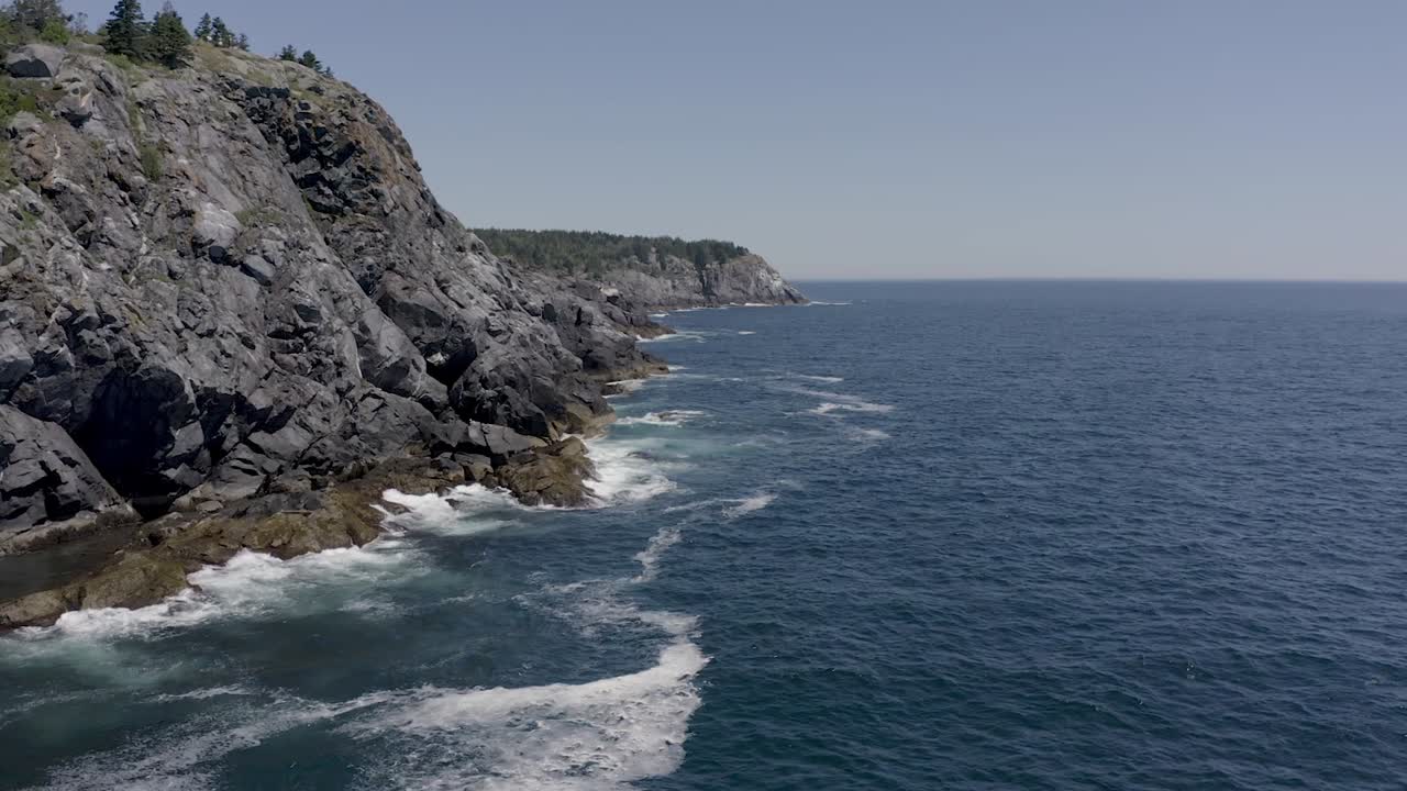 Drone flying around a cliff on the rocky coast in Maine to reveal more of the coastline with waves crashing into the rocks
