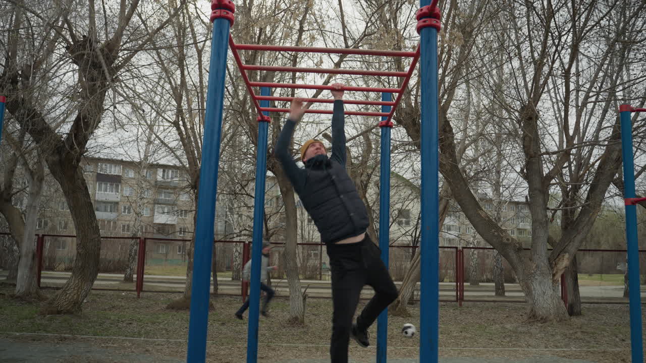 A coach jumps up and grabs a red iron bar, slowly moving across it with controlled strength, in the background, a boy plays football, and a residential building is visible across the road