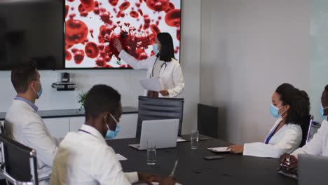 Mixed-race-female-doctor-giving-presentation-to-diverse-group-of-colleagues-wearing-masks