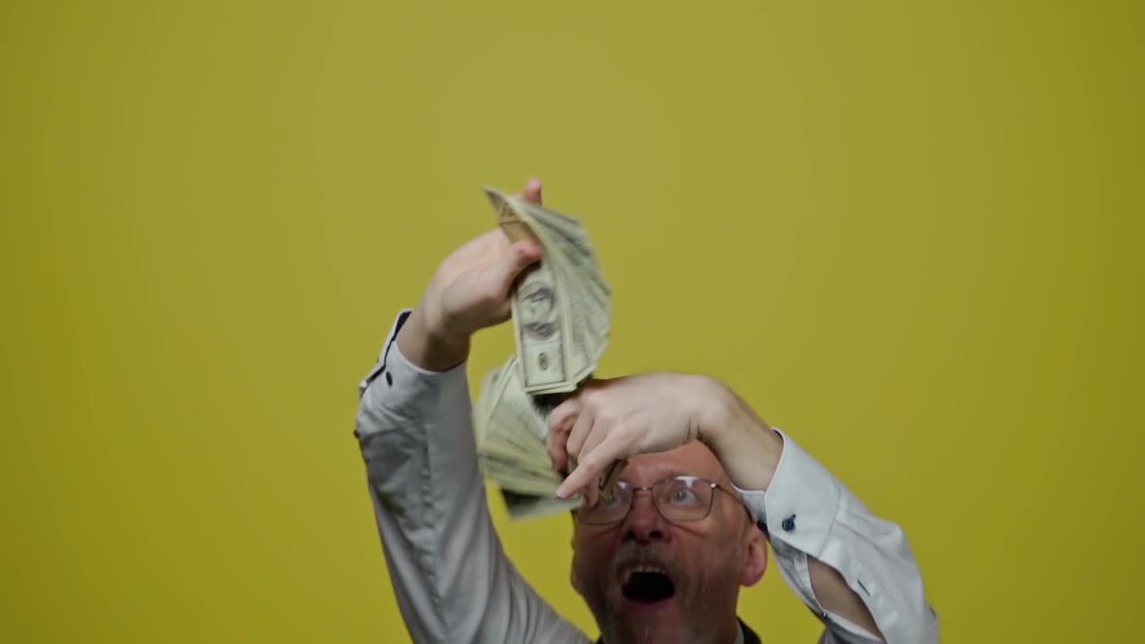 Good-looking joyful man with money on yellow background. Satisfied modern gray-bearded grandfather posing on camera with many dollar bills. Slow motion.