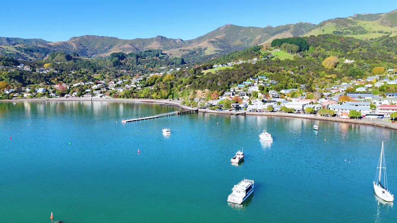 Aerial footage captures boats and lush hills surrounding Akaroa Harbour under clear skies, showcasing serene coastal beauty