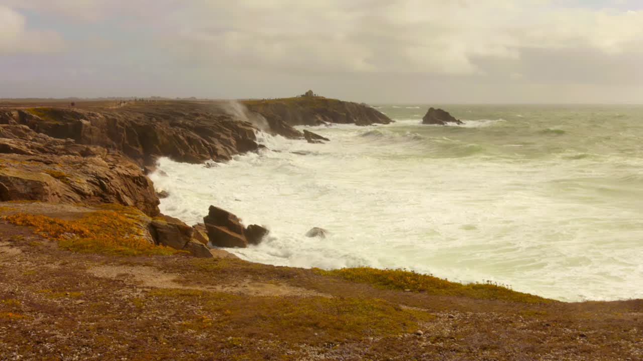 Dramatic wide shot of the rugged coastline of Brittany, France. Powerful, choppy Atlantic Ocean waves crash and foam against the dark cliffs and rocky shore of the Quiberon Peninsula