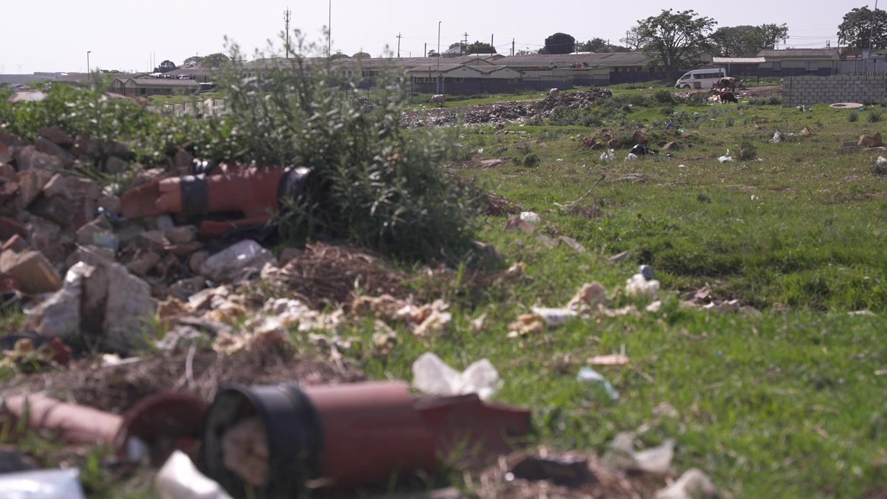 Rubble, plastic and construction debris litter a grassy field near low township buildings in bright midday light.