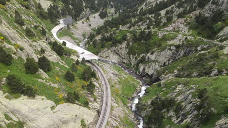 Toma-Aérea-De-Un-Paisaje-Montañoso-En-El-Que-Las-Vías-Del-Tren-Corren-A-Lo-Largo-De-Un-Rico-Río-En-Los-Pirineos