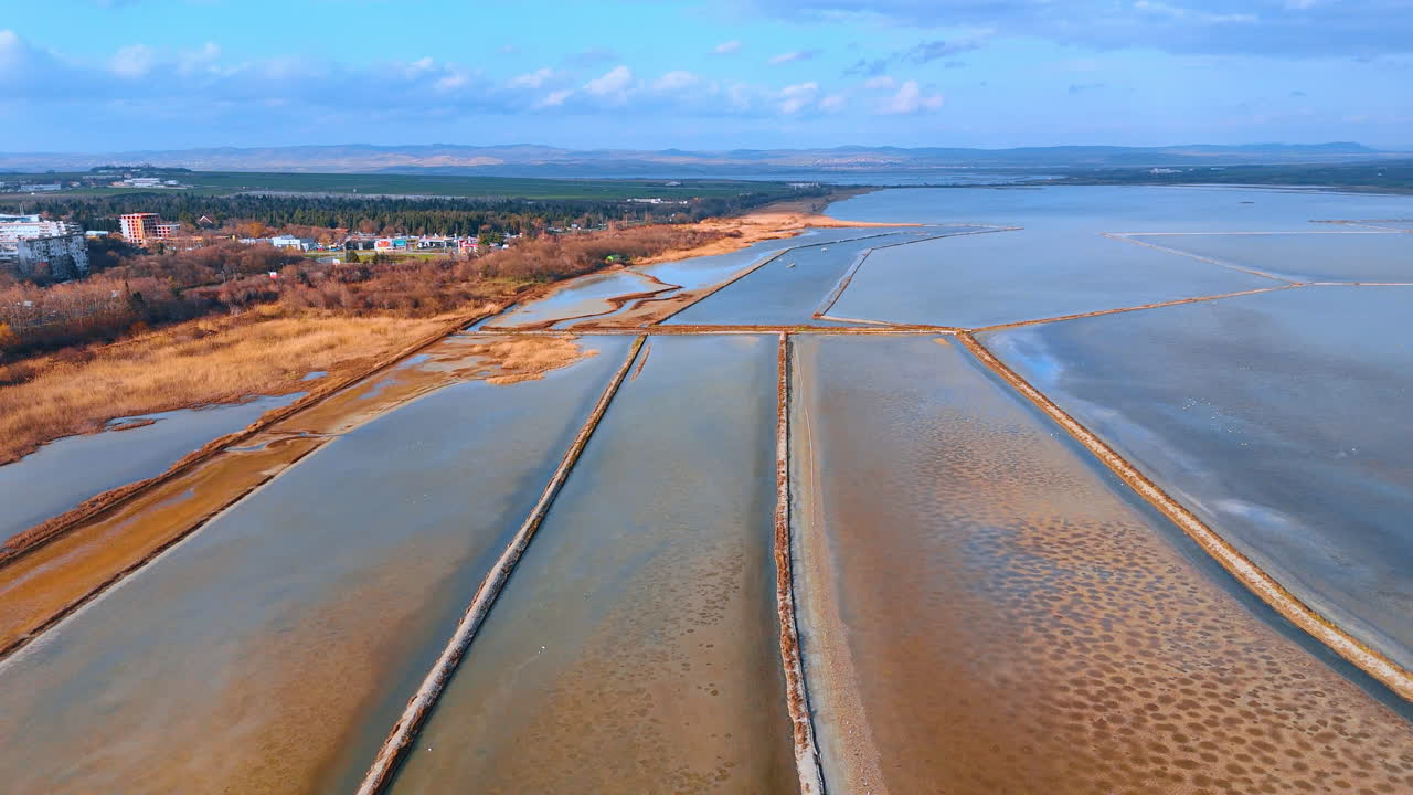 Luftfoto af saltpander og kystvegetation. Bredt dronebillede, der viser saltudvindingssted nær rørskov og bykant.