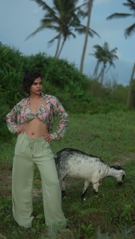woman stands outdoors with a goat grazing beside her against a lush tropical backdrop