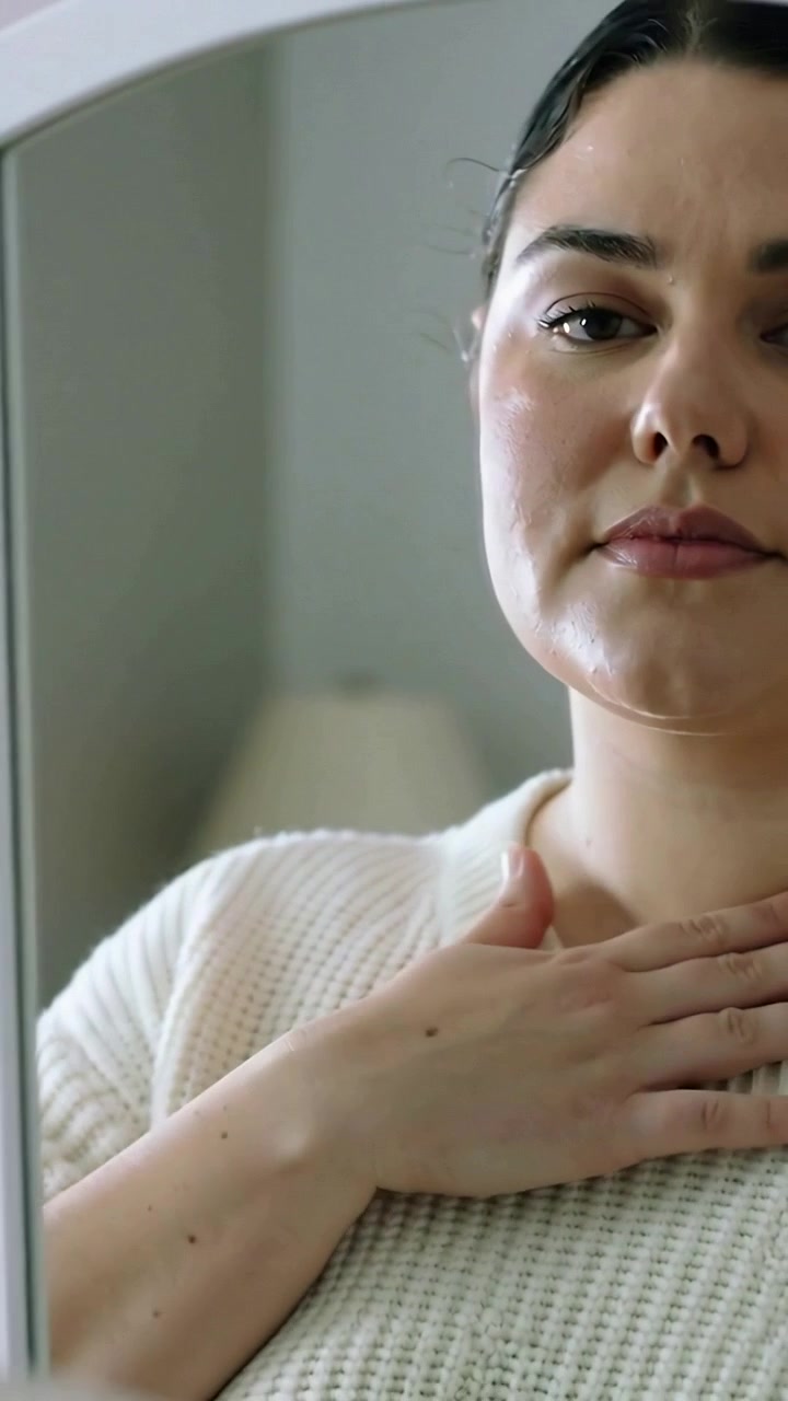 Video verticale: Toccandosi il collo, donna che si guarda allo specchio a casa, controllando il trucco e indossando una maglia bianca