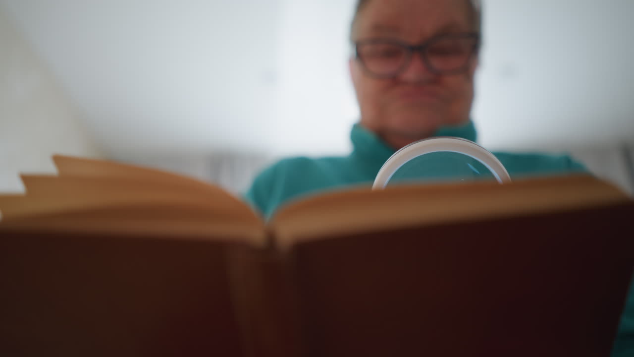 Elderly woman looking at book through magnifying glass, visibly focused on small print. She appears to be inspecting the text closely in her cozy bedroom, with a serious expression
