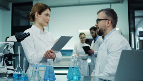 Medical-Laboratory-Male-And-Female-Scientists-Talking-About-Their-Researching-And-Discovery-While-Man-Sitting-At-The-Laptop-Computer-And-Woman-Standing-With-A-Folder