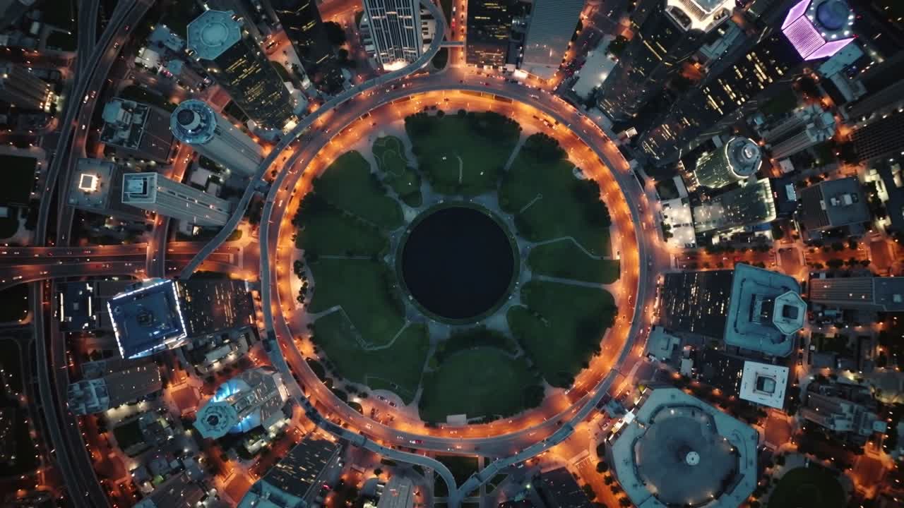 Dron ascendente y giratorio que revela un parque circular con estanque al anochecer, dejando a la vista las luces de la carretera de circunvalación