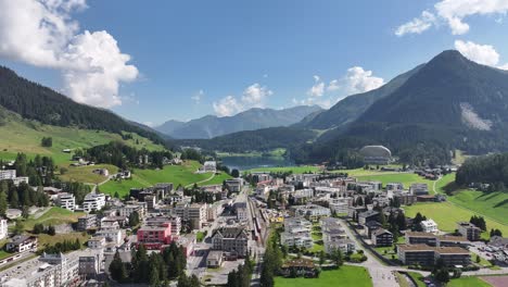 drone flies over town with architectural buildings, residential areas and lightly trafficked roads, set against tree filled mountains and lake in davos, kanton graubünden, switzerland