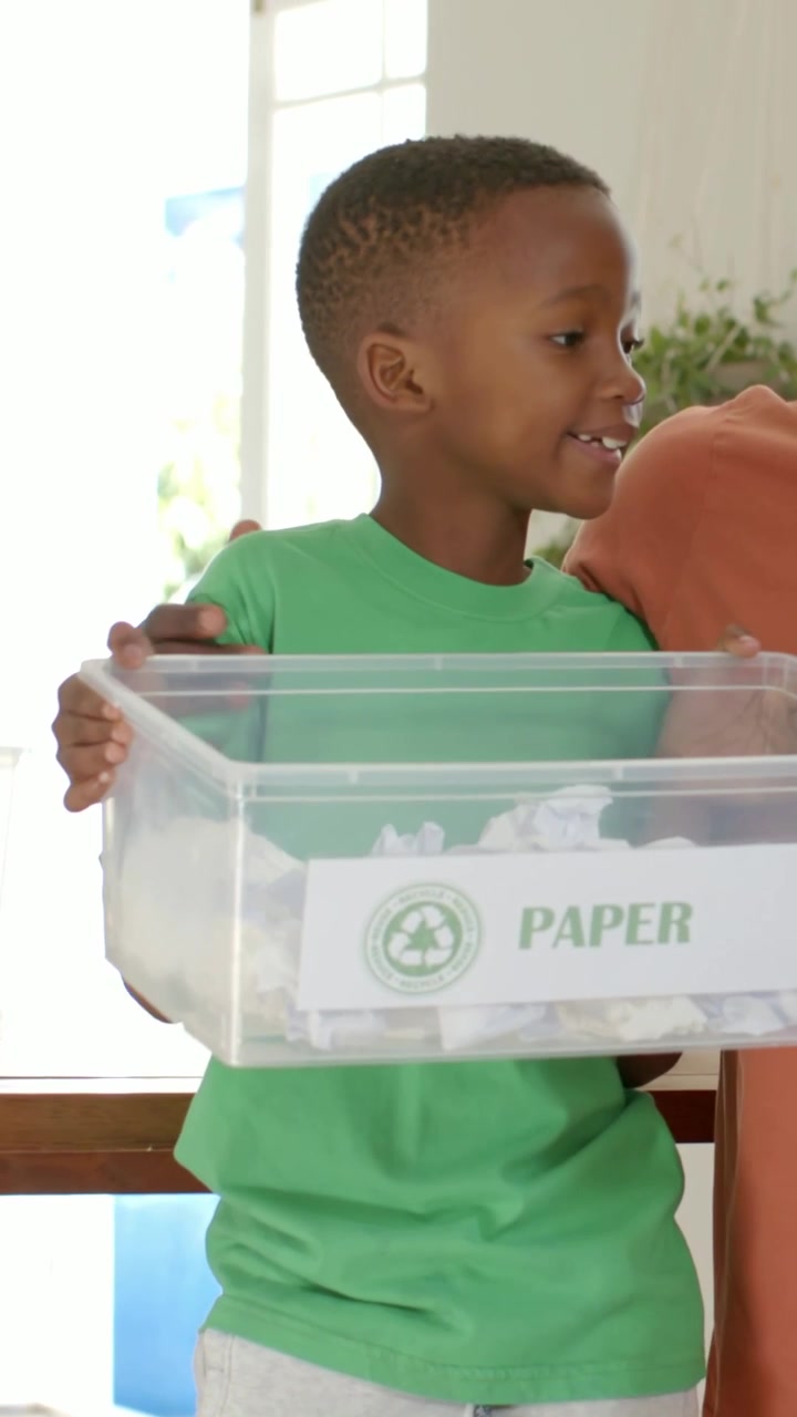 Vertical video: Arm reaching, parent guiding boy green tee holding PAPER bin at home to recycle