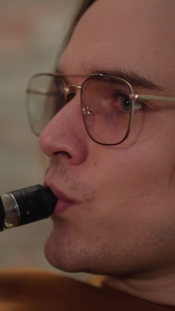joven elegante con gafas exhala humo blanco de un dispositivo de vape descansando en la sala de estar closeup cámara lenta. estilo de vida hipster y relajarse