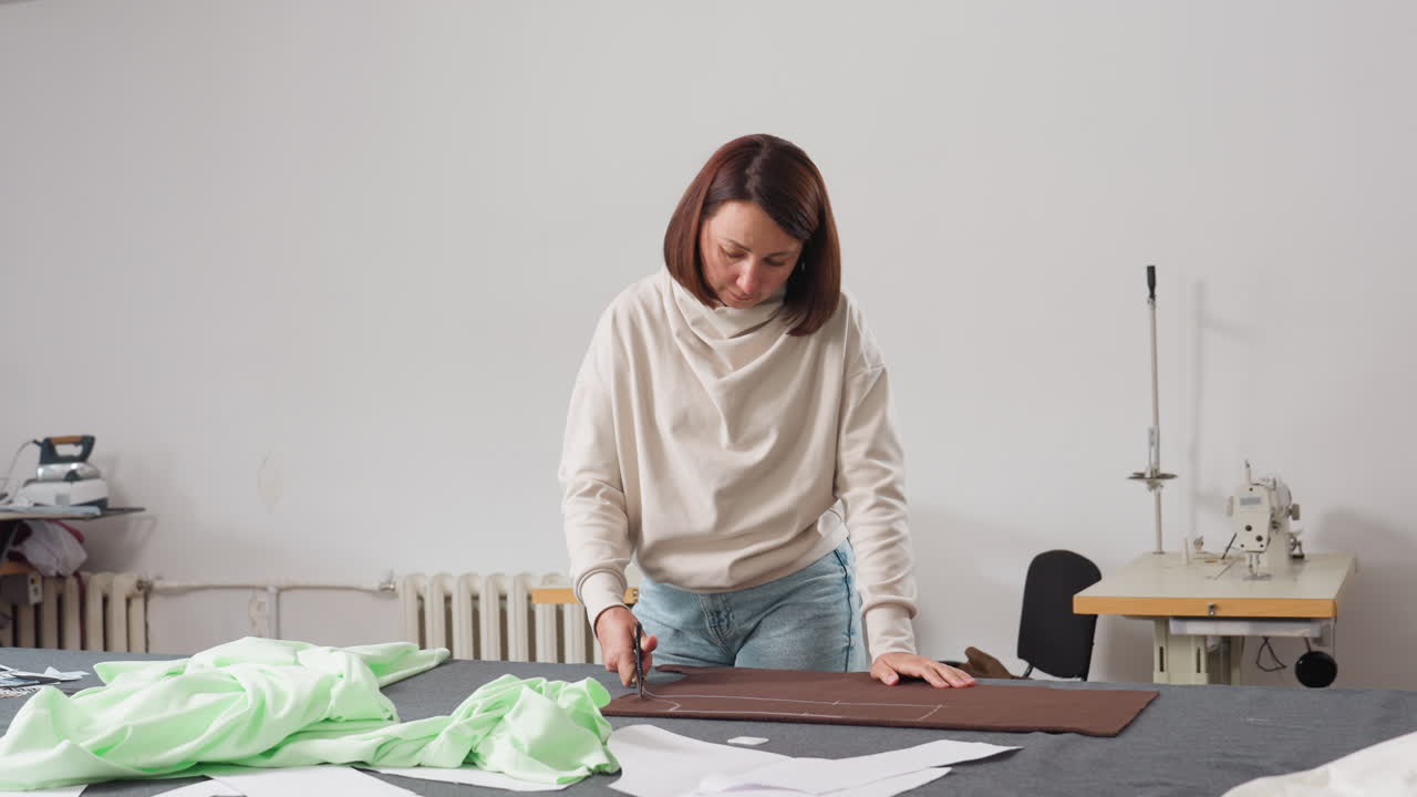 Seamstress cutting out design on brown fabric using large scissors on work table in sewing studio, surrounded by light green fabric, paper patterns, sewing tools, and sewing machine