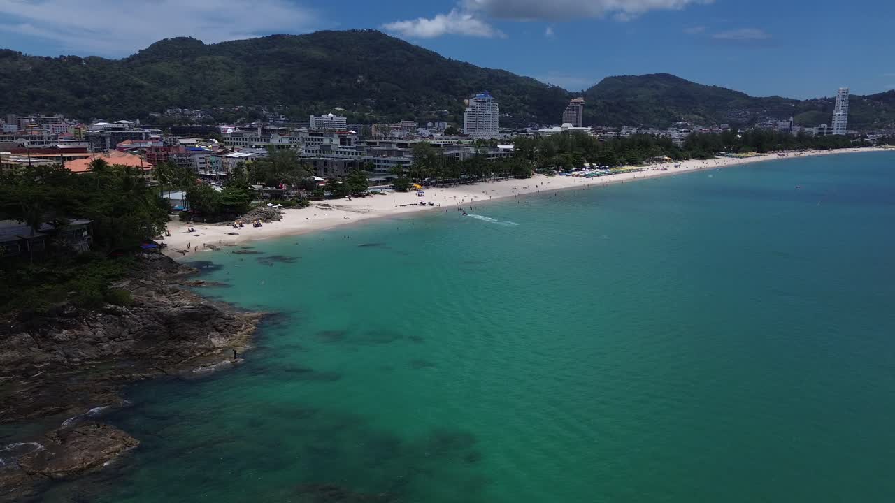 Aerial semi orbit of turquoise coastline and beachfront cityscape surrounded by lush green mountains in Phuket, Thailand