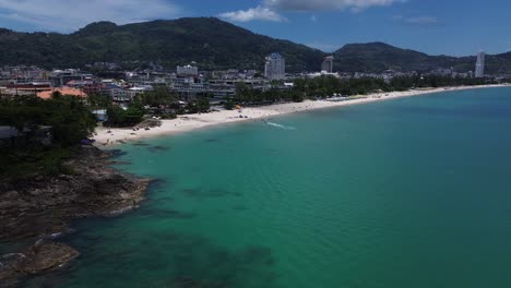 aerial semi orbit of turquoise coastline and beachfront cityscape surrounded by lush green mountains in phuket, thailand