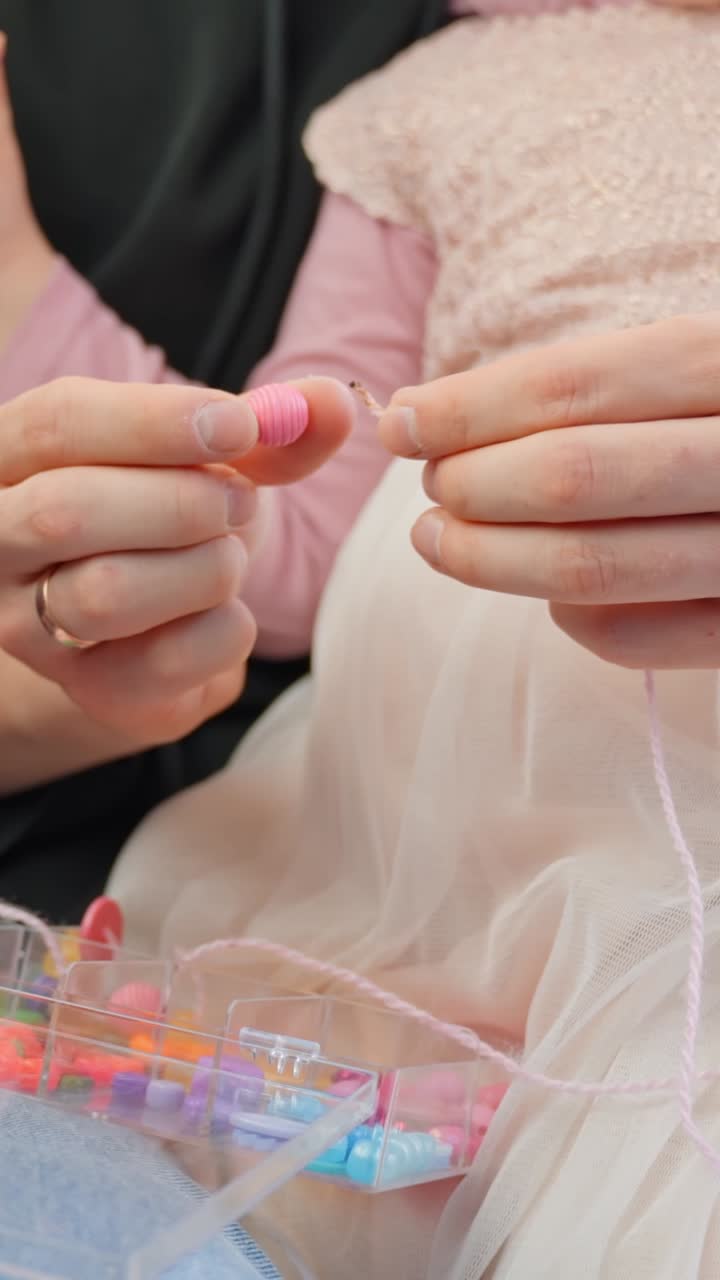 Hands arranging string beads working with young daughter to make necklace, soft indoor lighting, vertical