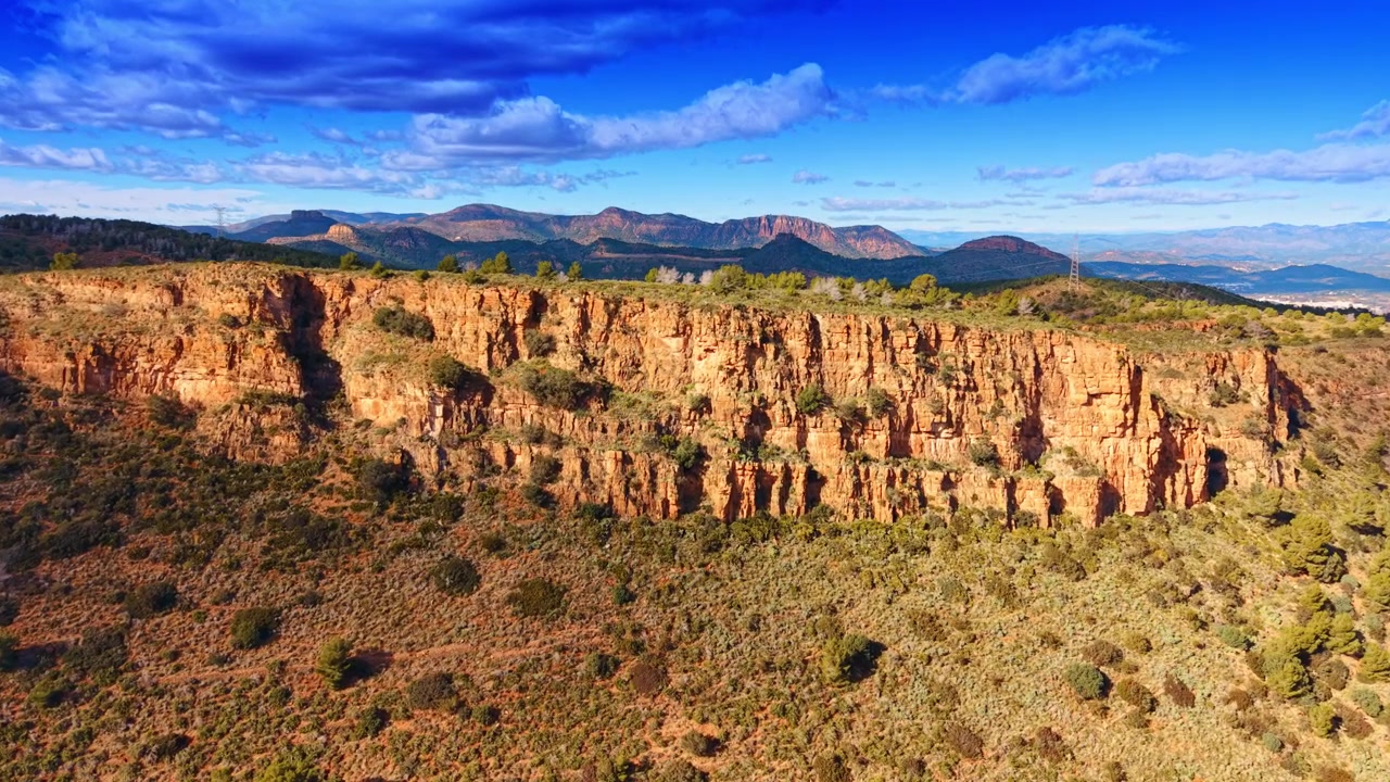 Paisagem cênica de falésias rochosas e terreno montanhoso sob nuvens. Vista panorâmica de falésias de calcário laranja, vegetação rasteira e cordilheiras montanhosas distantes sob céu azul.