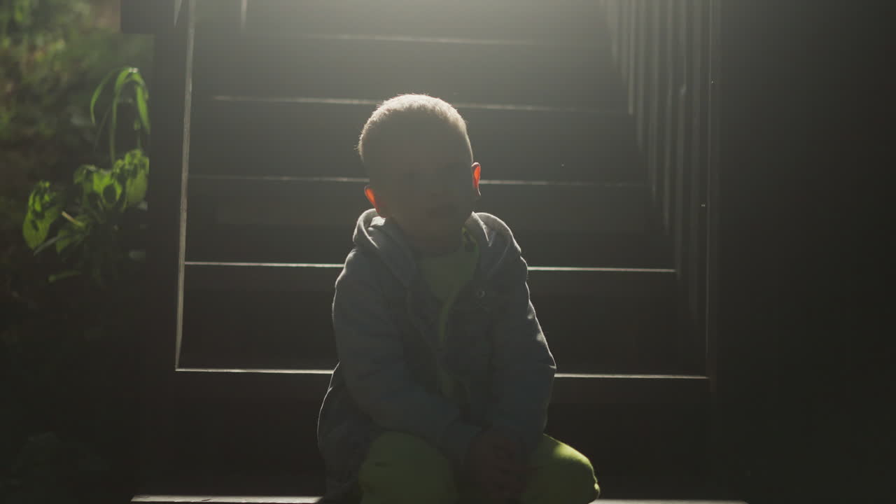 밤에 오두막 계단에 앉아있는 아이 실루: 작은 소년은 시골 집 입구의 계단에서 혼자 휴식을 취합니다. 버려진 아이는 주머니에 부모를 기다리고 있습니다.