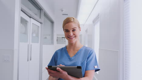 portrait of smiling female doctor wearing scrubs in hospital corridor using digital tablet