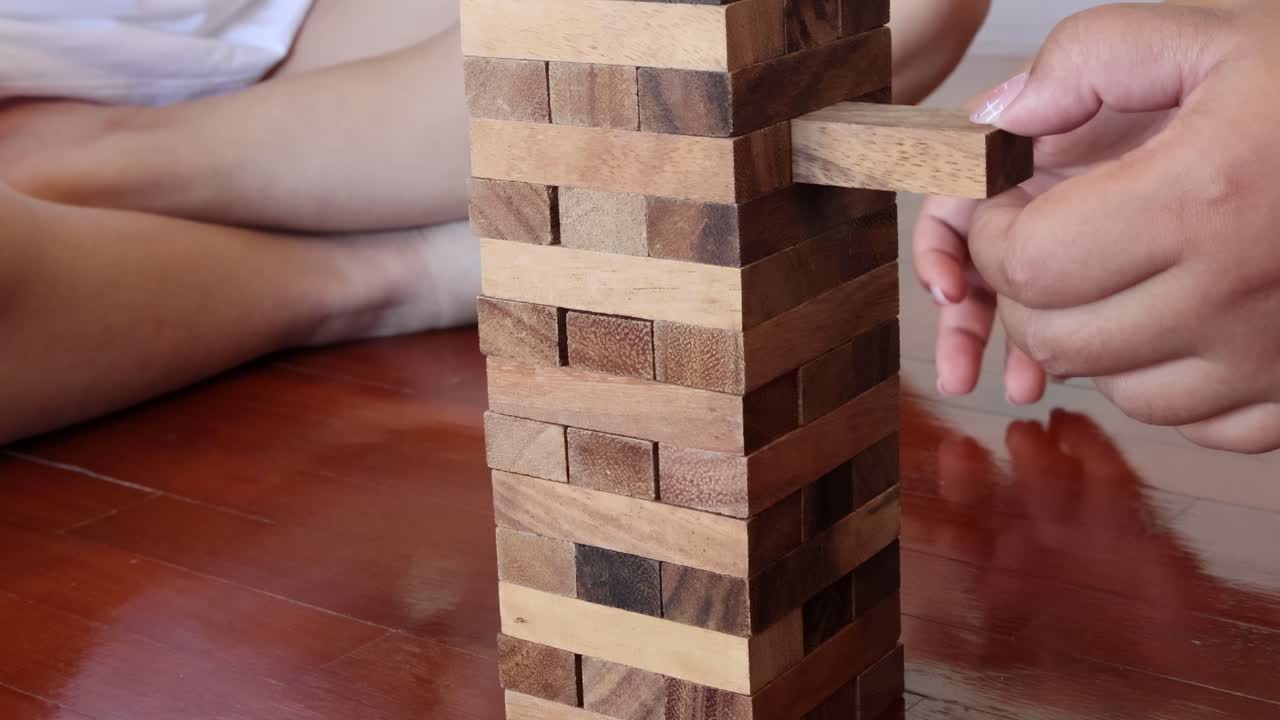 A detailed view of hands carefully extracting a block from a Jenga tower on a wooden floor.