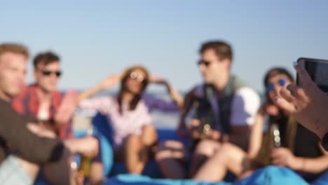 closeup view of a young hipster girl recording a video or taking pictures of group of friends sitting on easychairs on the beach, playing guitar and singing on a summer evening. slowmotion shot.