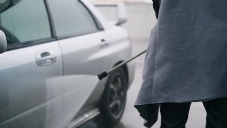 Young-woman-in-coat-washing-her-silver-sportcar-with-water-jet-on-self-service-carwash