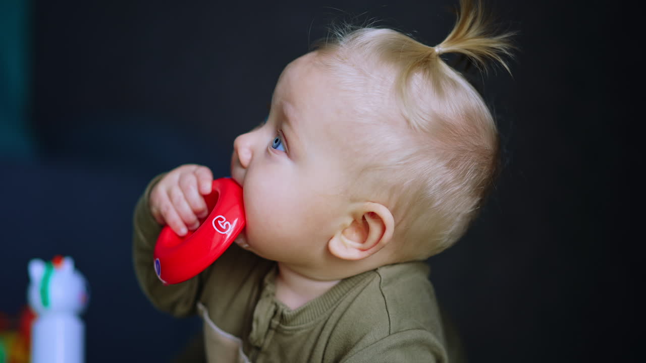 Beautiful baby with blond hair lies on his belly. Infant chews a red toy and lies down on his back. Close up.