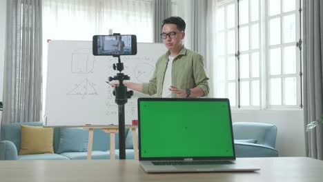asian male teacher with glasses and green screen laptop shoots video by smartphone camera while teaching math at home